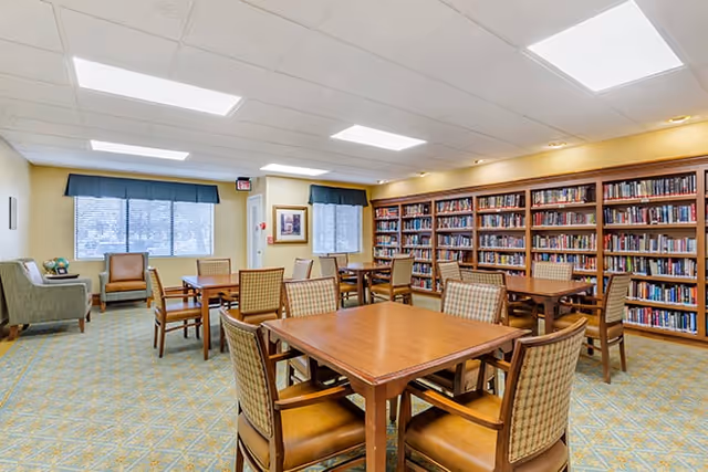 A well-lit room with multiple wooden tables and cushioned chairs arranged neatly. The room features a large bookshelf filled with books along one wall, two windows with blue valances, and a few armchairs near the windows. The carpet has a patterned design, and the ceiling has recessed lighting panels.