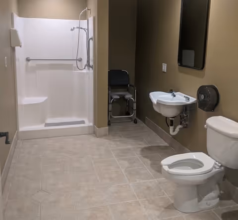 A clean and accessible bathroom with a white toilet, wall-mounted sink, large mirror, and a walk-in shower with grab bars. There is also a shower chair placed in the corner. The walls are painted beige and the floor is tiled.