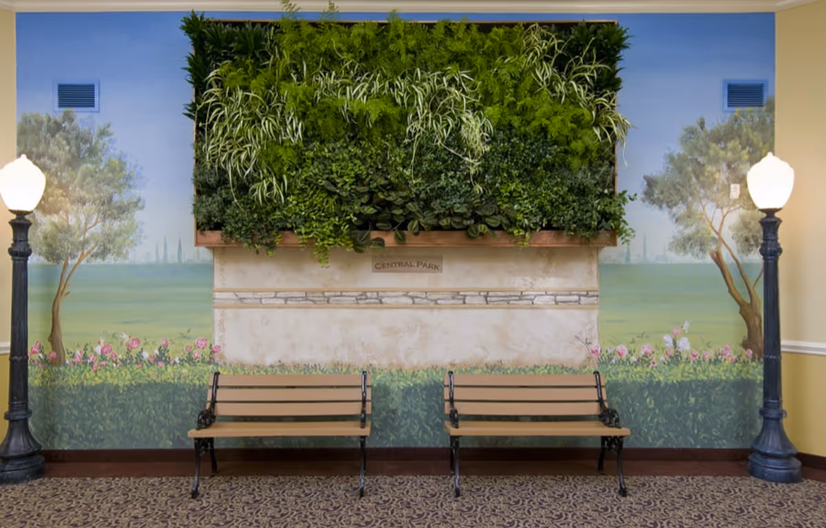 Indoor seating area with two wooden benches with black metal frames placed against a wall. The wall features a large vertical garden with various green plants and a mural depicting trees, flowers, and a landscape. Two vintage-style street lamps with white globes flank the benches. The floor is carpeted with a patterned design.
