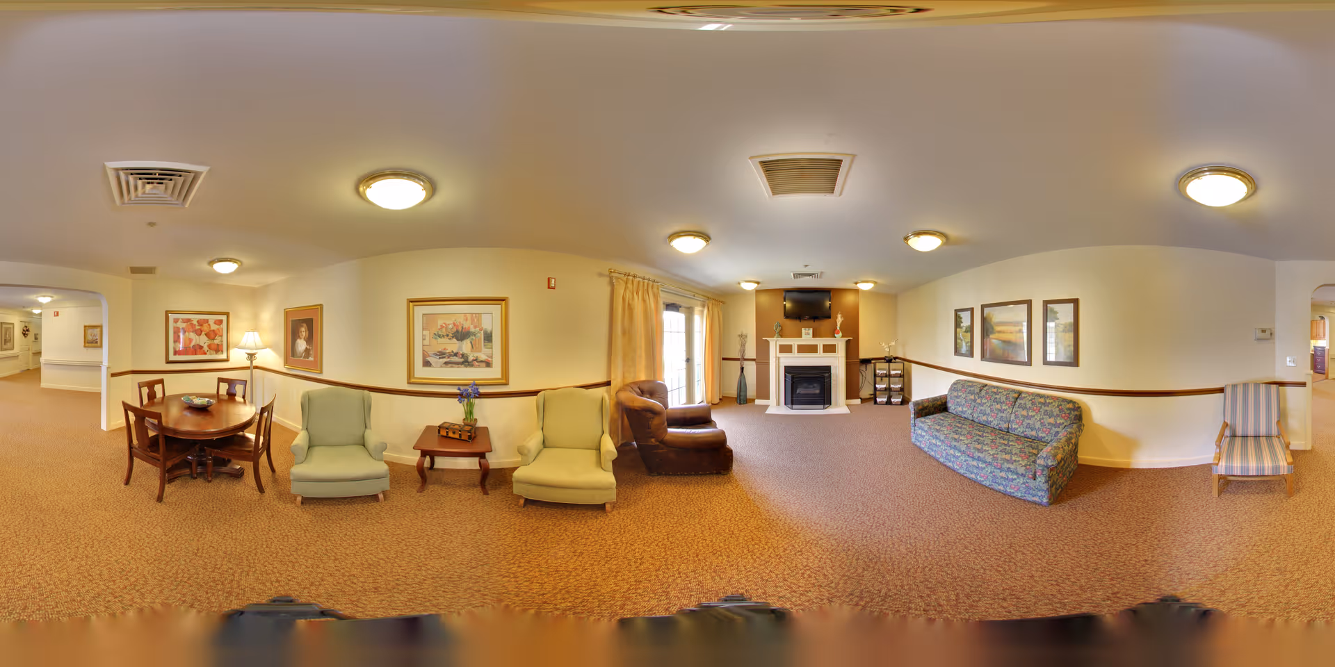 Panoramic view of a senior living common lounge with chairs, a sofa, a dining table, a fireplace with TV above, and framed artwork on the walls.