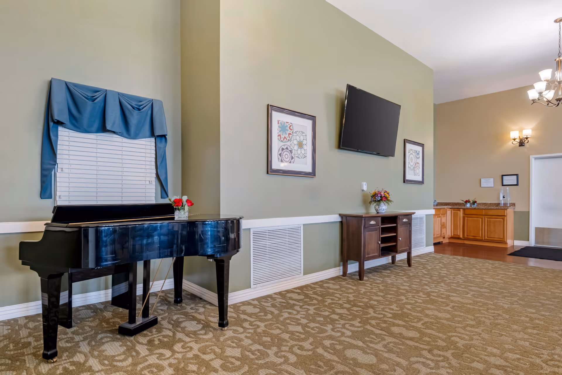 A senior living common area featuring a black grand piano, a wall-mounted TV above a wooden console with framed artwork, and a reception counter in the background.