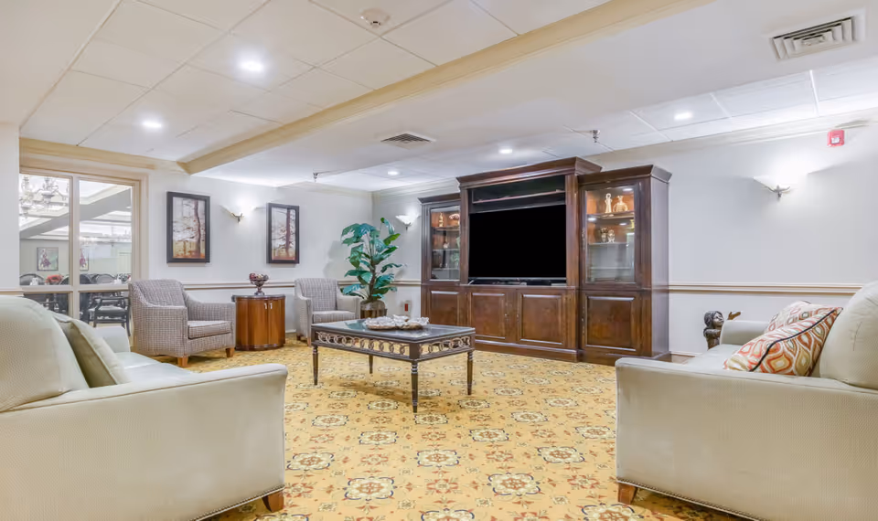 A senior community living room with beige sofas, two patterned armchairs, a wooden coffee table, and a large wooden entertainment center with a flat-screen TV. The room has patterned carpet, framed artwork on the walls, and a potted plant in the corner.