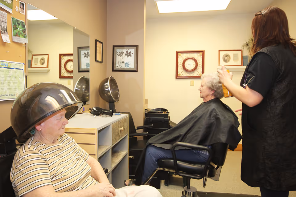 Two elderly women in a hair salon area of a senior living facility. One woman is sitting under a hair dryer wearing a striped shirt, while the other woman is seated in a salon chair with a black cape, having her hair styled by a hairdresser. The room has beige walls, framed pictures, a clock, and salon equipment.