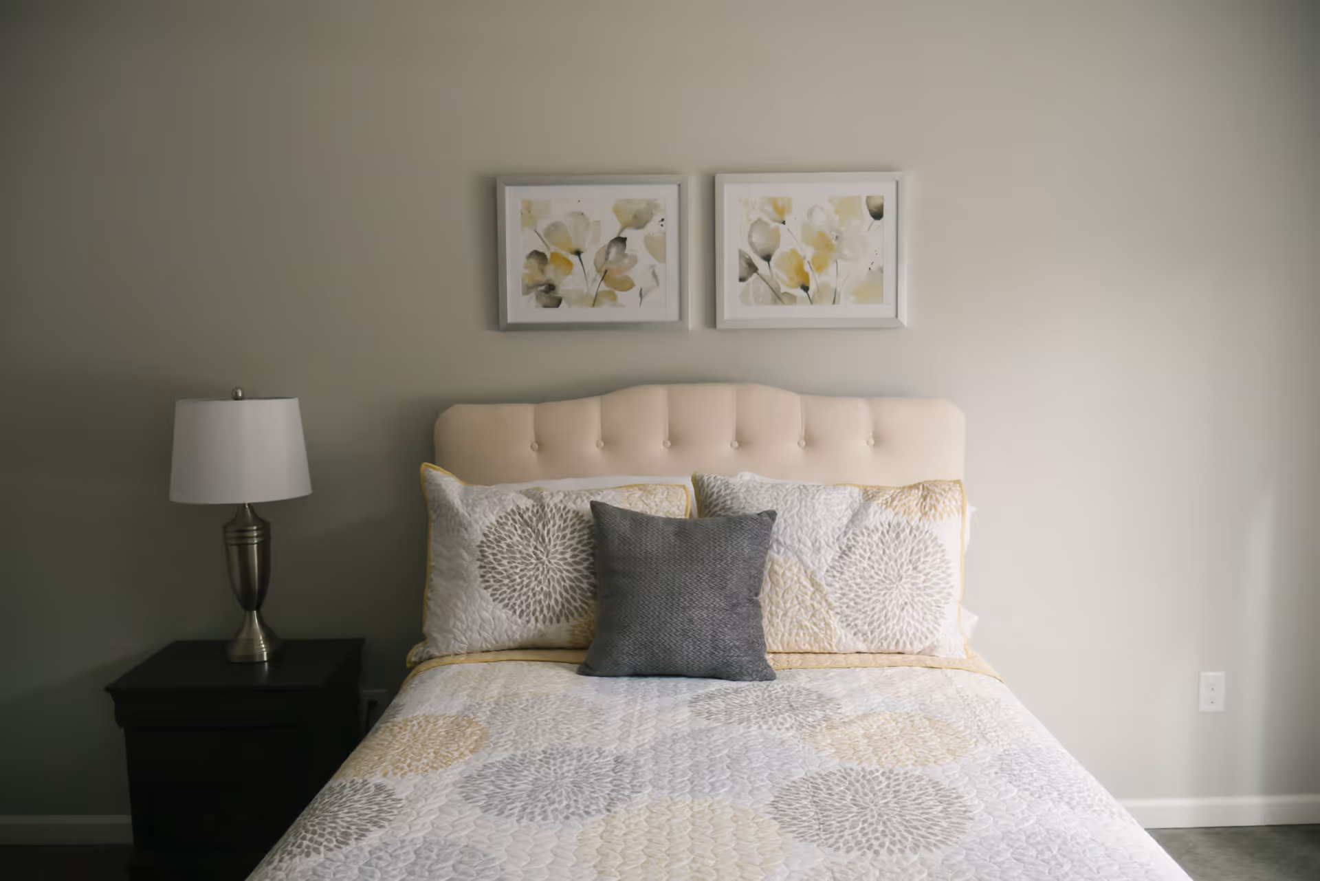 A neatly made bed with a beige tufted headboard, white and yellow floral patterned bedding, and a gray decorative pillow in the center. On the left side of the bed is a dark wooden nightstand with a silver lamp with a white lampshade. Above the bed are two framed floral paintings with yellow and gray tones. The walls are painted light gray.