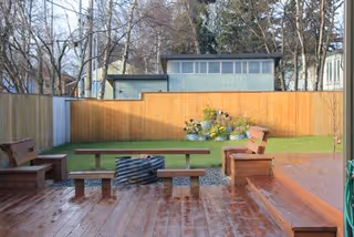 Outdoor seating area with wooden benches and a fire pit on a wooden deck, surrounded by a grassy yard and a wooden fence with trees in the background.