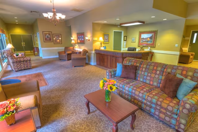 A spacious and warmly lit assisted living facility common area with patterned sofas and armchairs arranged around wooden coffee tables. The room features a reception desk with a chair behind it, framed artwork on the walls, and several lamps providing soft lighting. The carpeted floor and yellow walls create a cozy atmosphere.