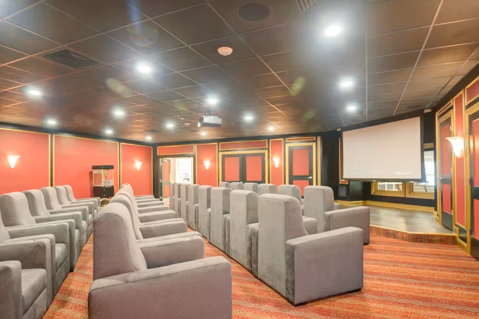 A small theater room with rows of gray upholstered armchairs facing a large projector screen. The walls are painted red with gold trim, and the ceiling is black with recessed lighting. There is a popcorn machine in the back corner of the room.