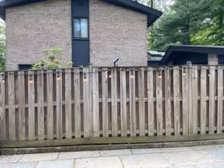 A wooden fence in front of a brick building with a tall narrow window. There are trees and greenery visible behind the fence.