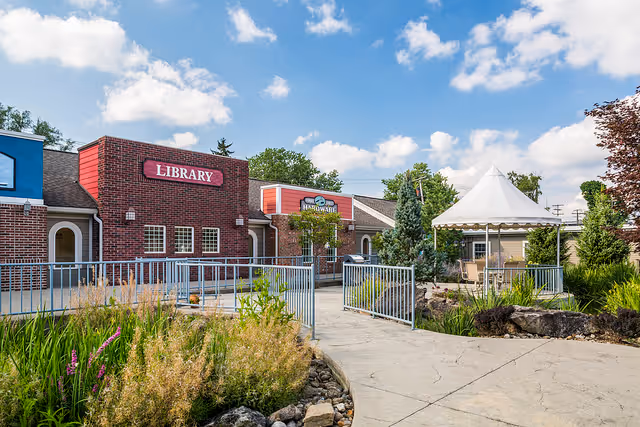Outdoor courtyard featuring a brick building labeled "Library", a paved walkway, a gazebo, and landscaped greenery under a blue sky.