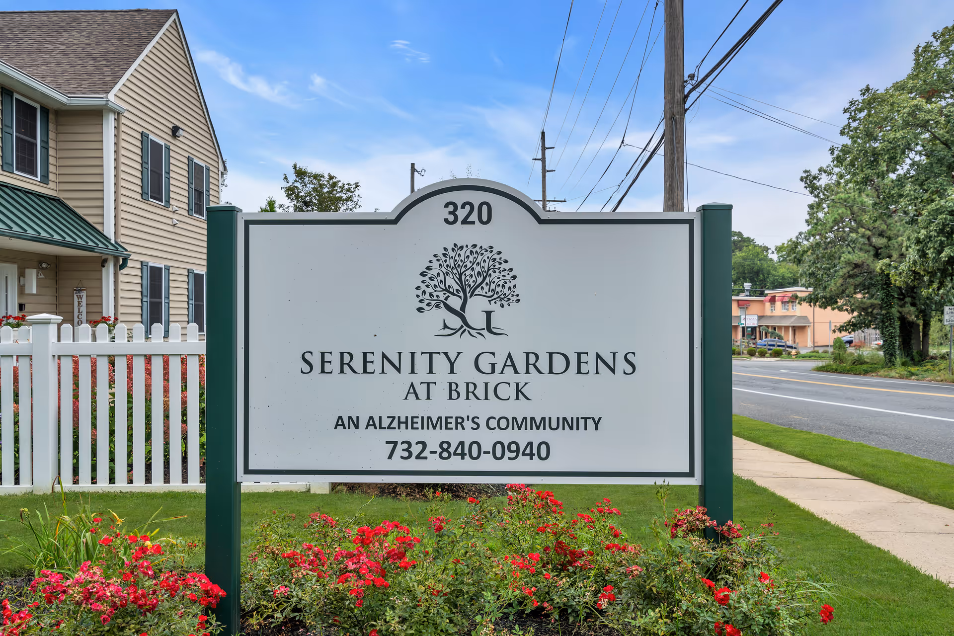 Outdoor view of a sign for Serenity Gardens at Brick, an Alzheimer's community, with the address number 320 and phone number 732-840-0940. The sign is surrounded by red flowers and greenery, with a beige building and a white picket fence in the background, along with a road and trees.