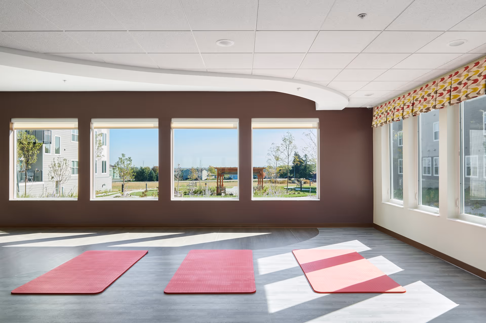 A bright room with large windows letting in natural light, showing an outdoor garden area. Inside the room, three red yoga mats are laid out on a light gray floor, suggesting a space for exercise or relaxation.