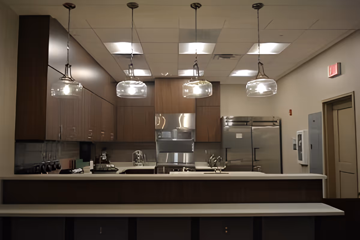 Modern kitchen area with wooden cabinets, stainless steel refrigerator and oven, a white countertop island with four hanging glass pendant lights above, and a door on the right side of the room.