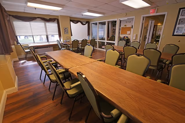 A well-lit dining room with wooden floors and multiple tables arranged in rows, surrounded by green cushioned chairs. Large windows with brown curtains allow natural light to fill the room. The walls are painted yellow, and there are framed pictures and a bulletin board on the walls.