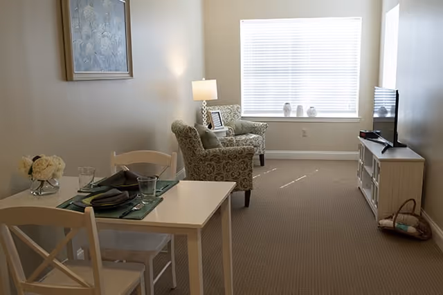 A cozy living space with a small dining table set for two in the foreground, two patterned armchairs near a window with white blinds, a floor lamp, and a TV on a white stand against the wall. The room has beige carpeting and neutral-colored walls with a framed artwork hanging.