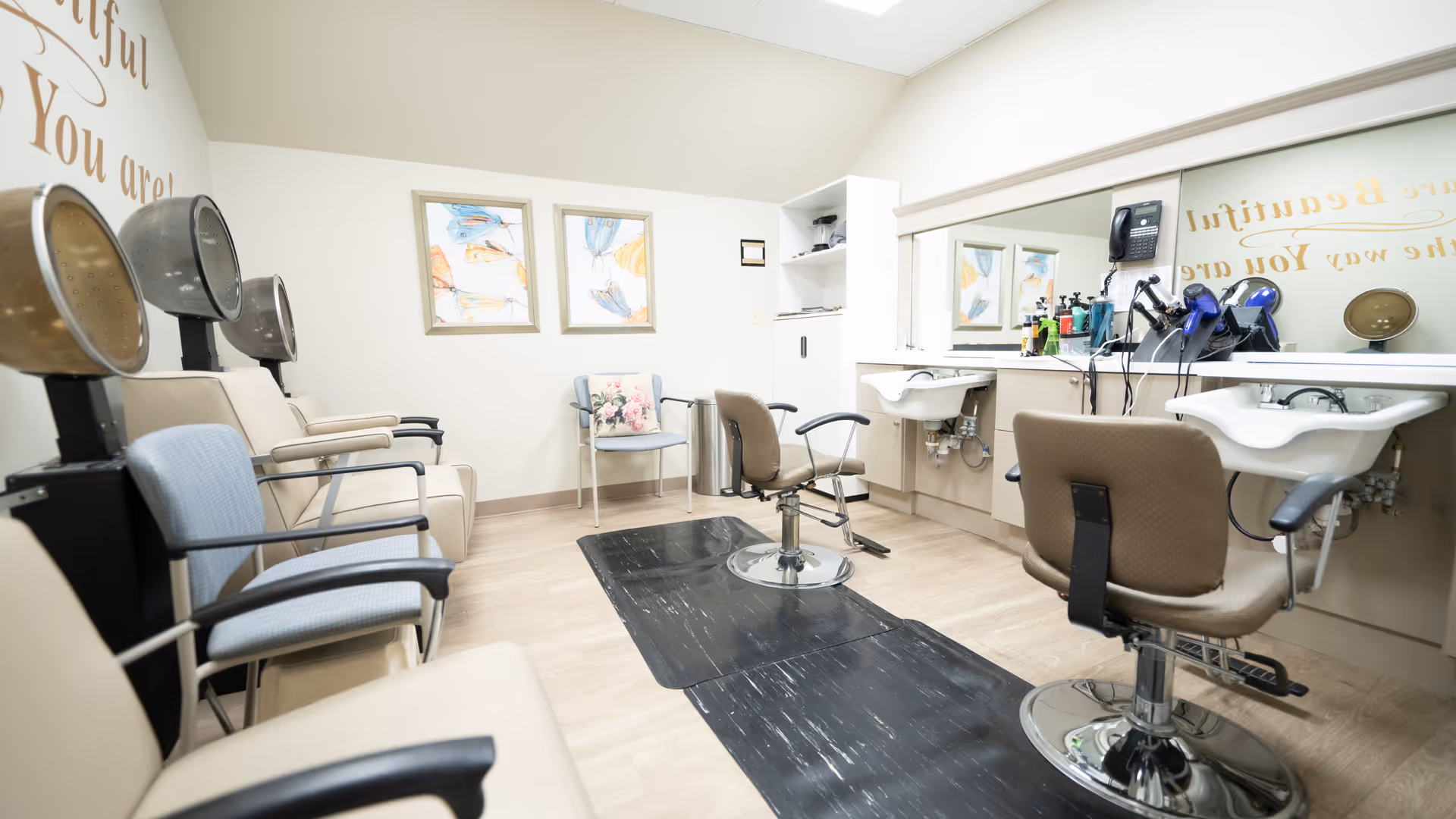 Interior view of a hair salon area in a senior living facility with salon chairs, hair dryers, sinks, and a large mirror. The room has light-colored walls, framed artwork, and motivational wall text partially visible.