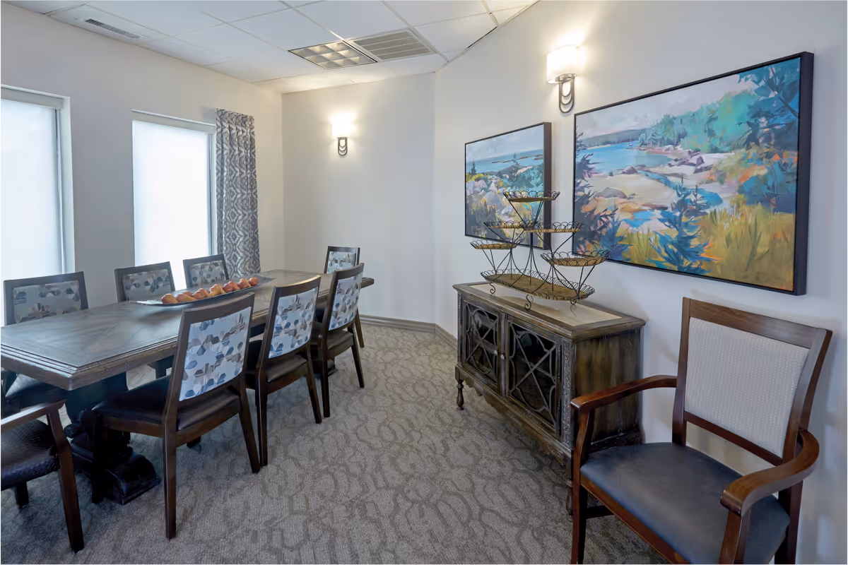 A dining room with a long wooden table surrounded by chairs, a sideboard holding decorative tiered trays, and colorful landscape paintings on the wall.