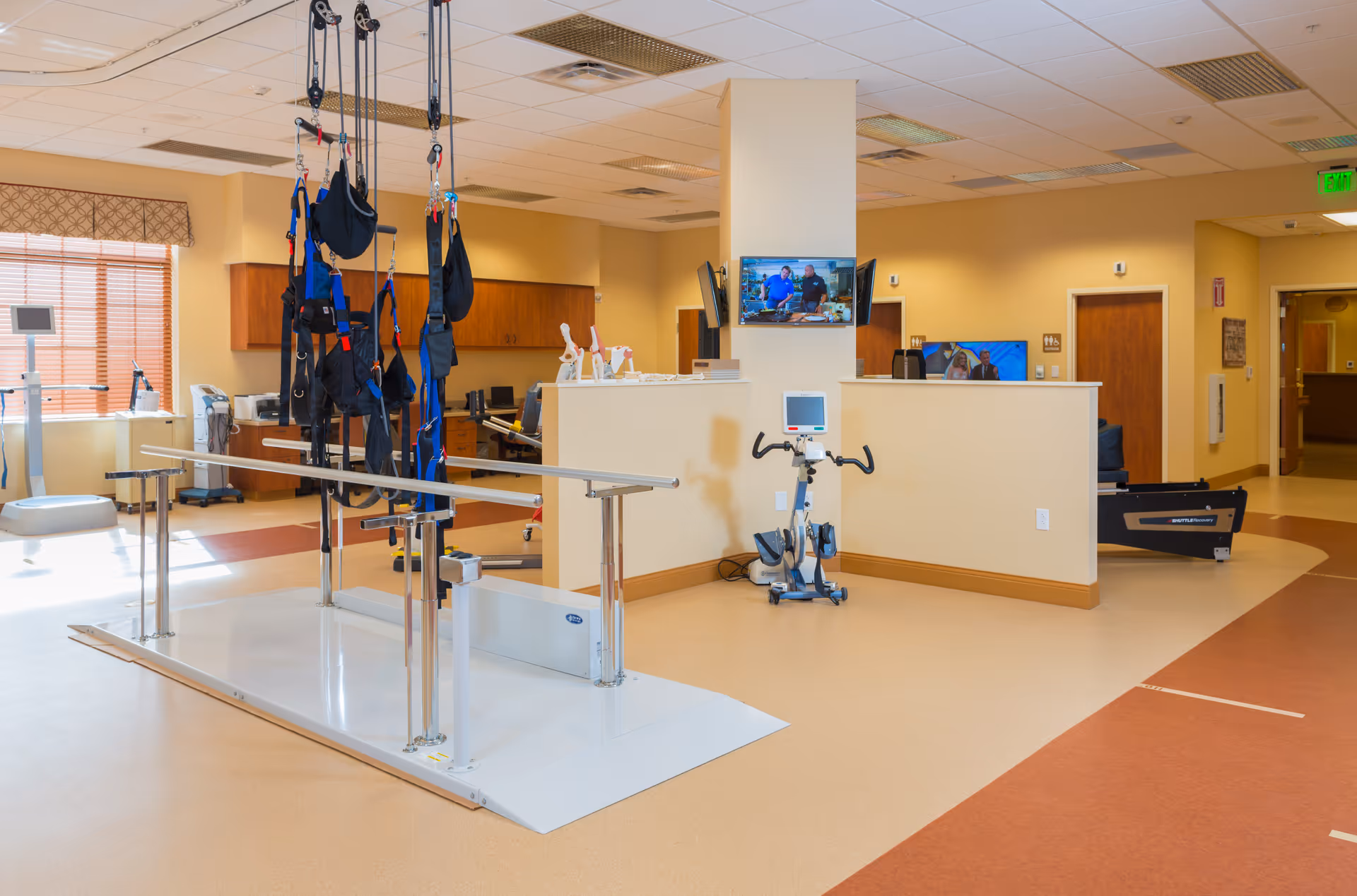 A spacious physical therapy room with parallel bars and harnesses for gait training, exercise equipment including a stationary bike, and a reception desk with two mounted TVs in the background.