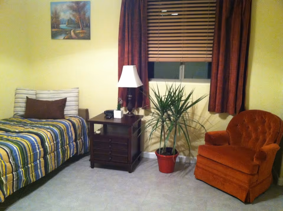 A cozy bedroom with a single bed covered in a striped yellow, green, and blue comforter, a brown pillow, a wooden nightstand with a lamp, a potted plant in a red pot, an orange upholstered armchair, and a window with closed blinds and red curtains.
