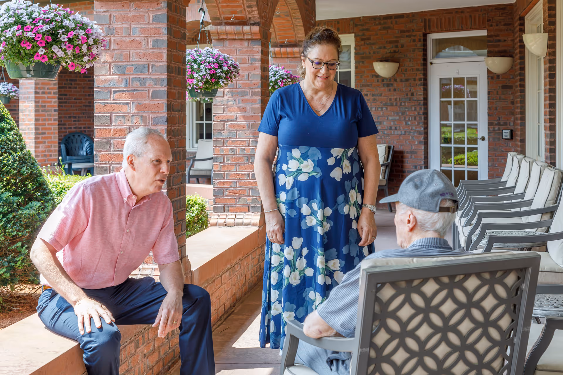Three elderly people socializing on a covered outdoor patio with brick pillars and hanging flower baskets. One man in a pink shirt is sitting on a low brick wall, a woman in a blue floral dress is standing, and another man wearing a gray cap is seated in a chair facing them. The patio has multiple chairs lined up along the wall and a glass door in the background.