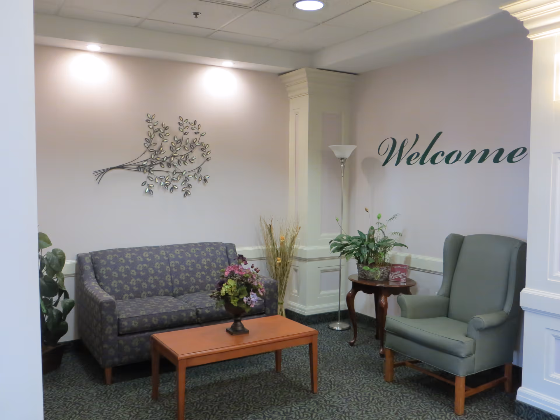 A cozy sitting area in a senior living community featuring a patterned loveseat, a wooden coffee table with a floral arrangement, a green upholstered armchair, a small round side table with a potted plant, a floor lamp, and a wall decoration of a metal branch with leaves. The word 'Welcome' is written on the wall in elegant script.