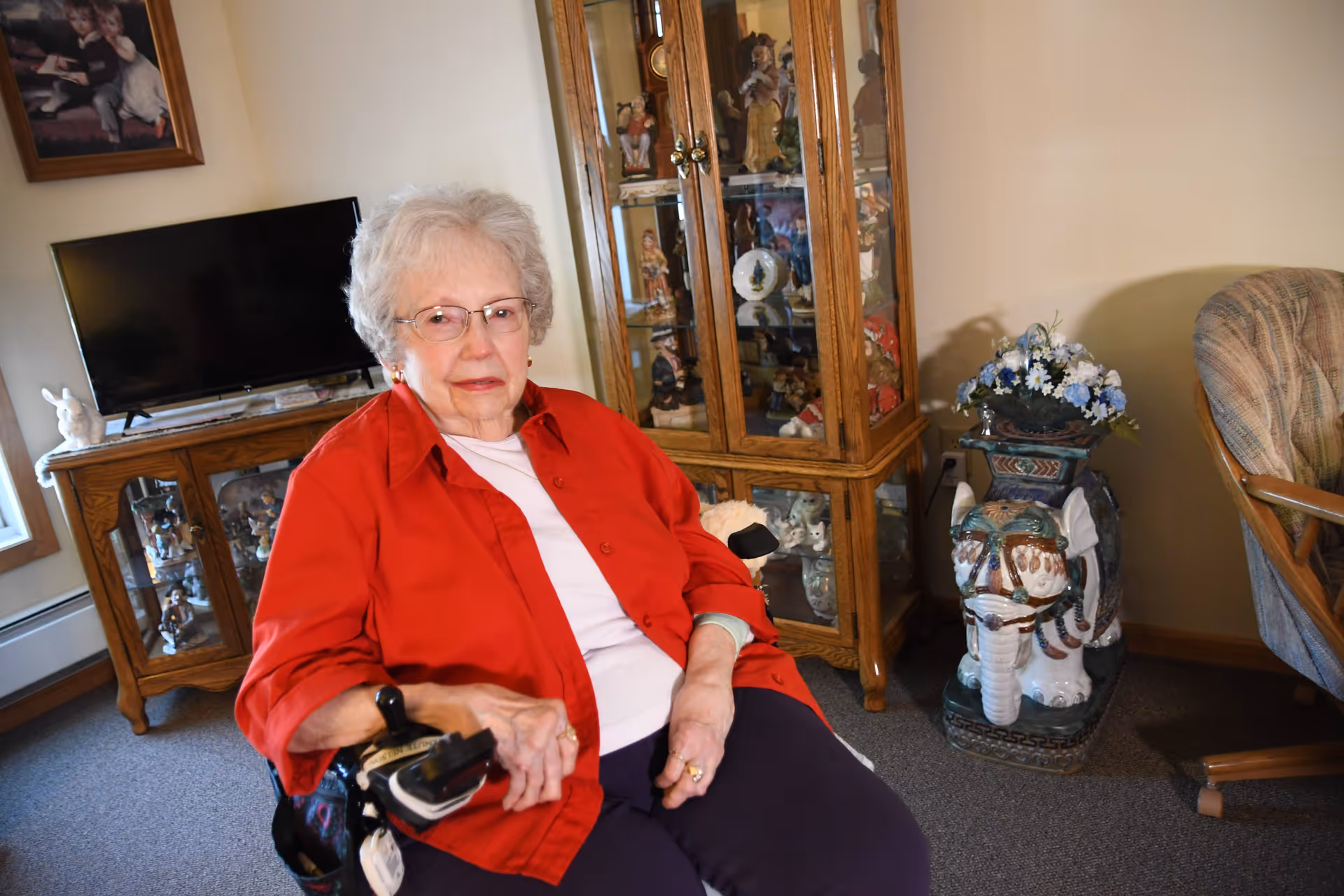 An elderly woman with white hair and glasses wearing a red shirt and white top sits in a wheelchair in a living room. Behind her is a wooden cabinet with glass doors displaying figurines, a TV on a wooden stand, a framed picture on the wall, a decorative ceramic elephant planter with flowers, and a cushioned armchair.