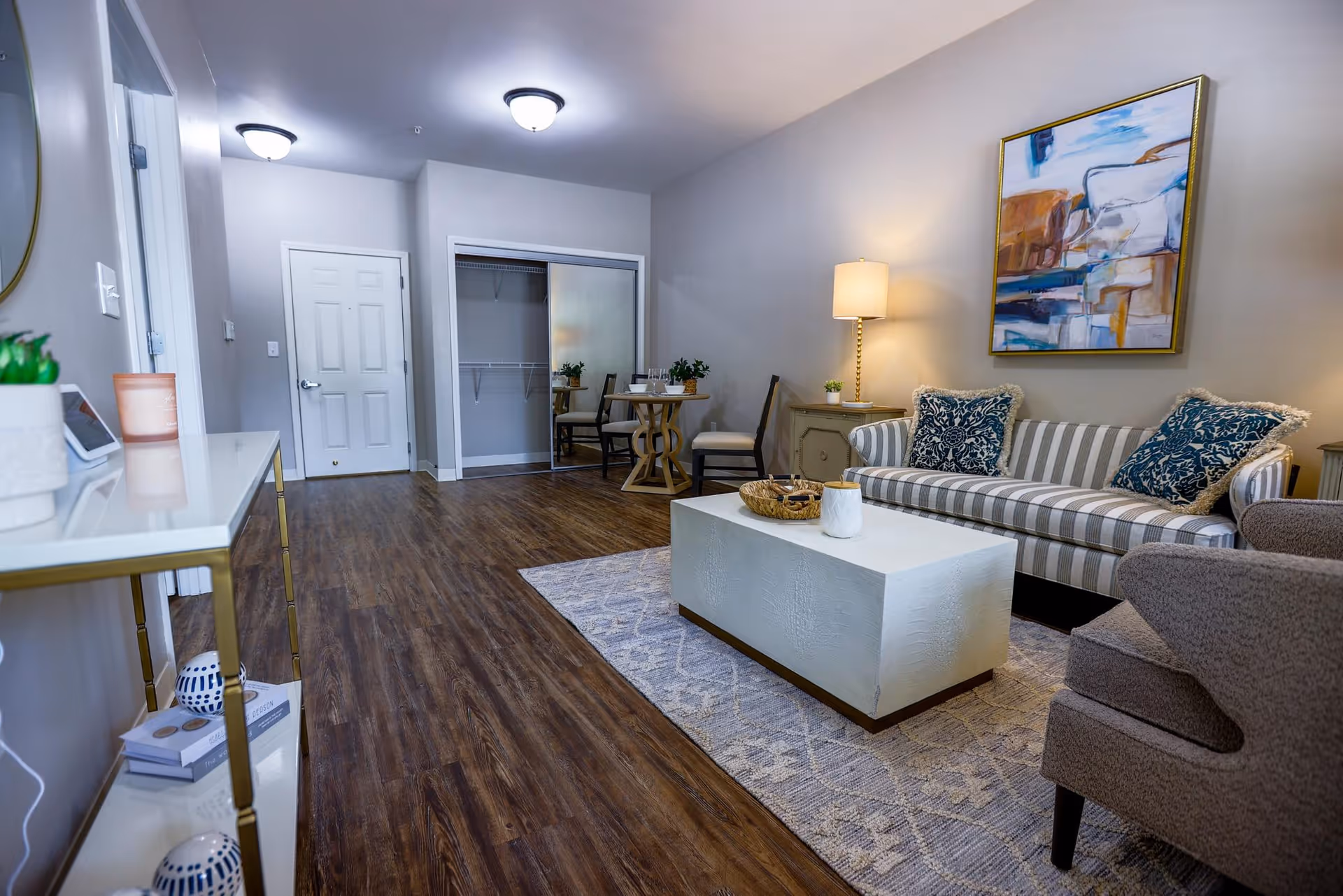 A cozy living room with a striped sofa adorned with two patterned blue pillows, a white rectangular coffee table on a patterned rug, a gray armchair, a side table with a lamp, and a small dining table with two chairs near a closet with mirrored sliding doors. The room has wood flooring and neutral-colored walls with a colorful abstract painting hanging above the sofa.