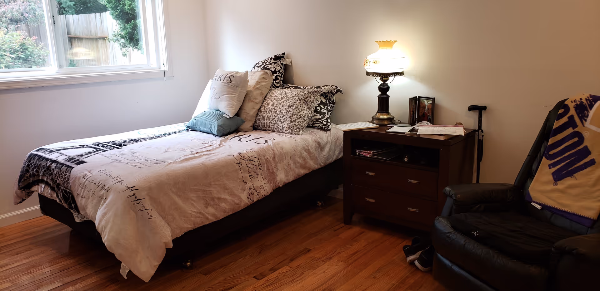 A cozy bedroom with a single bed covered in a light-colored comforter and multiple pillows. Next to the bed is a wooden nightstand with a vintage-style lamp, some books, and papers. A black leather recliner chair with a blanket draped over the back is positioned near the nightstand. A window on the left side lets in natural light, and the floor is wooden.