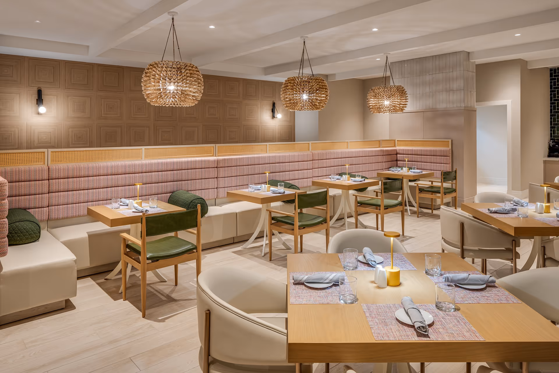 A modern dining room with wooden tables set with plates, glasses, and napkins. The seating includes cushioned benches along the wall with pink and beige upholstery, green chairs, and cream-colored armchairs. Three decorative pendant lights hang from the ceiling, and the walls feature a textured wooden panel design and neutral paint.