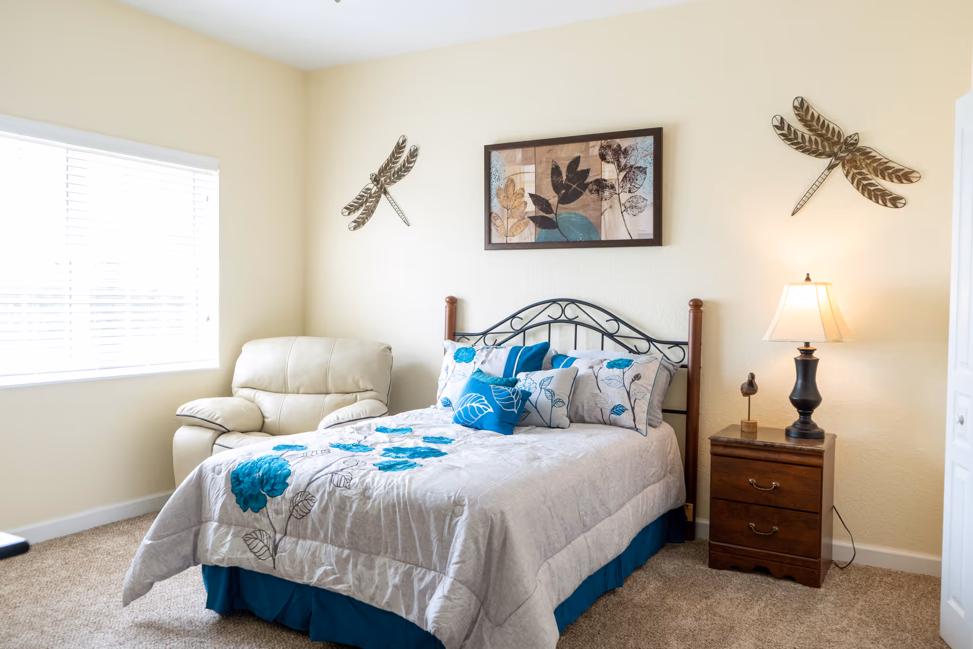 A bright bedroom with a bed featuring a floral comforter and multiple pillows in shades of blue and beige. There is a beige armchair next to a window with white blinds on the left side. On the right side, there is a wooden nightstand with a black lamp and a small decorative bird sculpture. The walls are light-colored and decorated with two metal dragonfly wall hangings and a framed artwork of leaves above the bed.