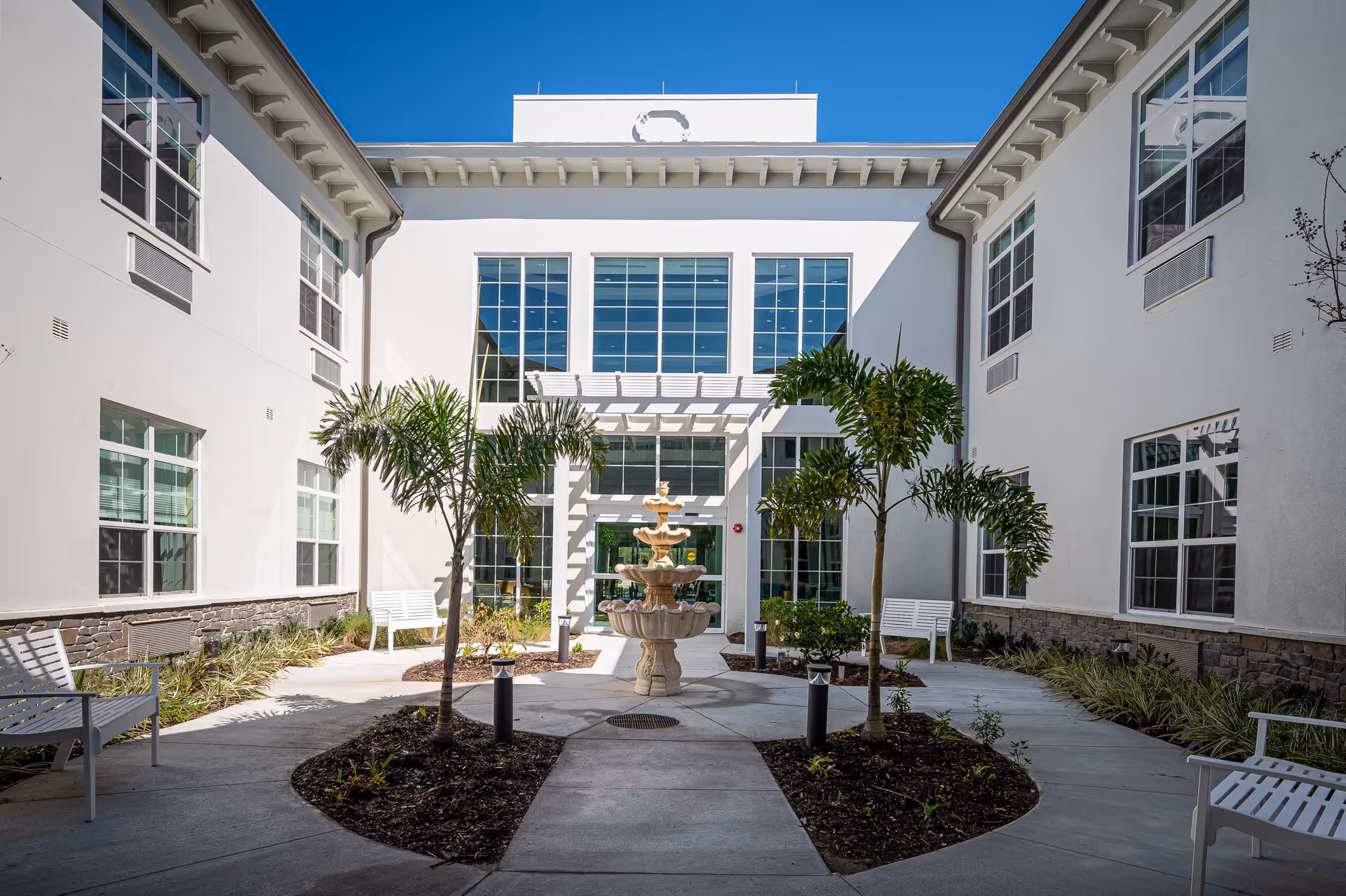 Outdoor courtyard area of a senior living facility with a central multi-tiered stone fountain surrounded by small palm trees and landscaped garden beds. White benches are placed along the concrete pathways, and the building's white exterior walls with large windows frame the courtyard under a clear blue sky.