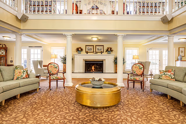 A spacious and well-lit living room area with two green sofas adorned with floral cushions, two wooden chairs with floral upholstery, a large round gold coffee table with a decorative bowl, and a fireplace with a fire burning. The room features white columns, patterned carpet, and a balcony railing above with framed photos and decorations.