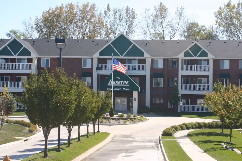 Exterior view of a three-story residential building with balconies, green triangular roof accents, and an American flag flying in front. The entrance has a green awning with the text 'The Reserve' and the address '2727 82nd Place'. Trees and well-maintained landscaping line the driveway leading to the entrance.