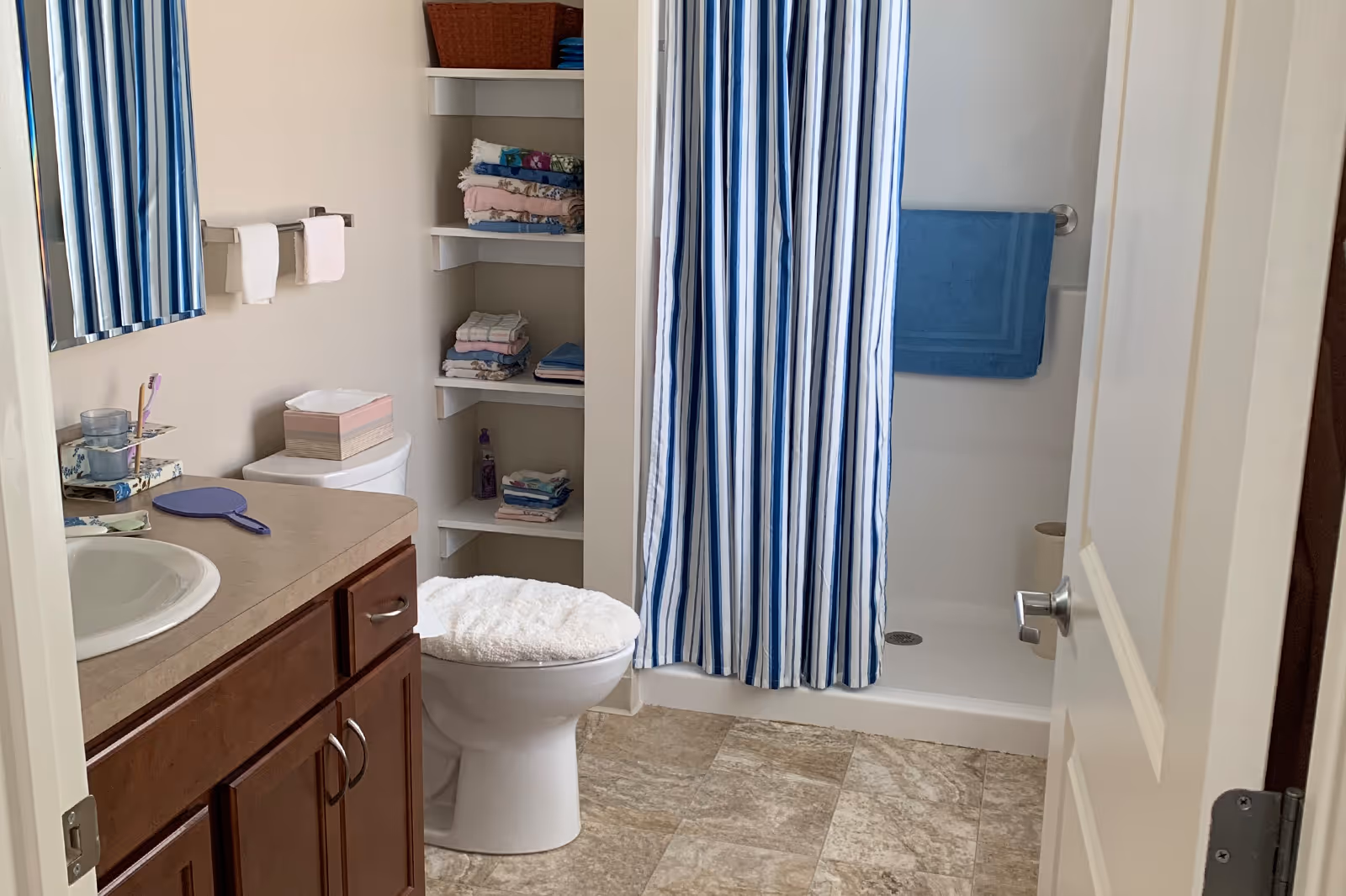 Bathroom with a vanity sink, toilet with a fuzzy seat cover, open shelving of folded towels, and a shower with a blue-and-white striped curtain.