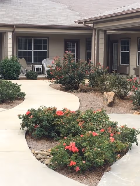 Curved concrete walkway surrounded by landscaped garden beds with flowering bushes leading to the entrance of a single-story building with beige siding and multiple windows. There are outdoor chairs and a small table on the porch area.