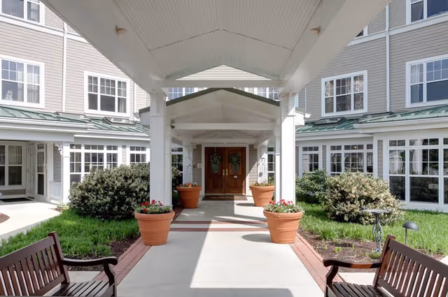 Covered entrance walkway to a building with white pillars and a peaked roof, flanked by large flower pots with red and white flowers. There are two wooden benches on either side of the walkway and bushes and grass along the sides. The building has multiple windows and a double wooden door at the entrance.