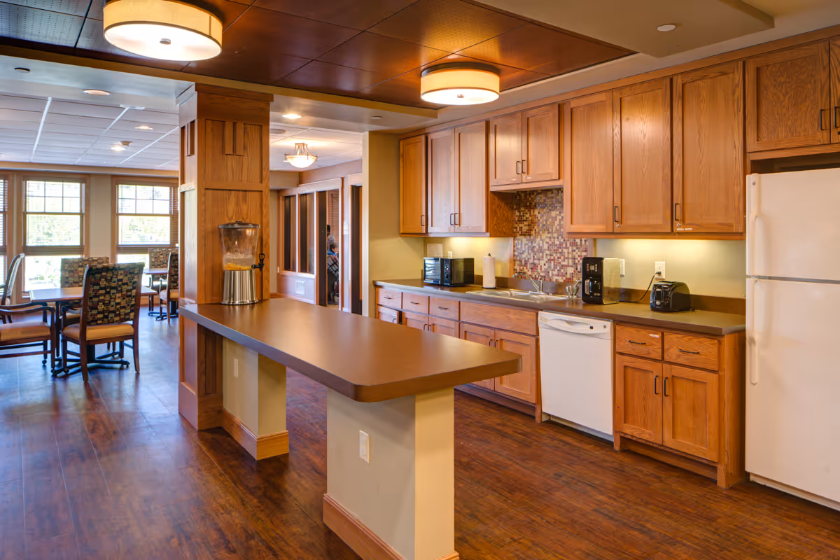 Interior view of a senior living facility kitchen area with wooden cabinets, a white refrigerator, dishwasher, coffee maker, toaster, and a water dispenser on a long counter. In the background, there is a dining area with tables and chairs near large windows letting in natural light.