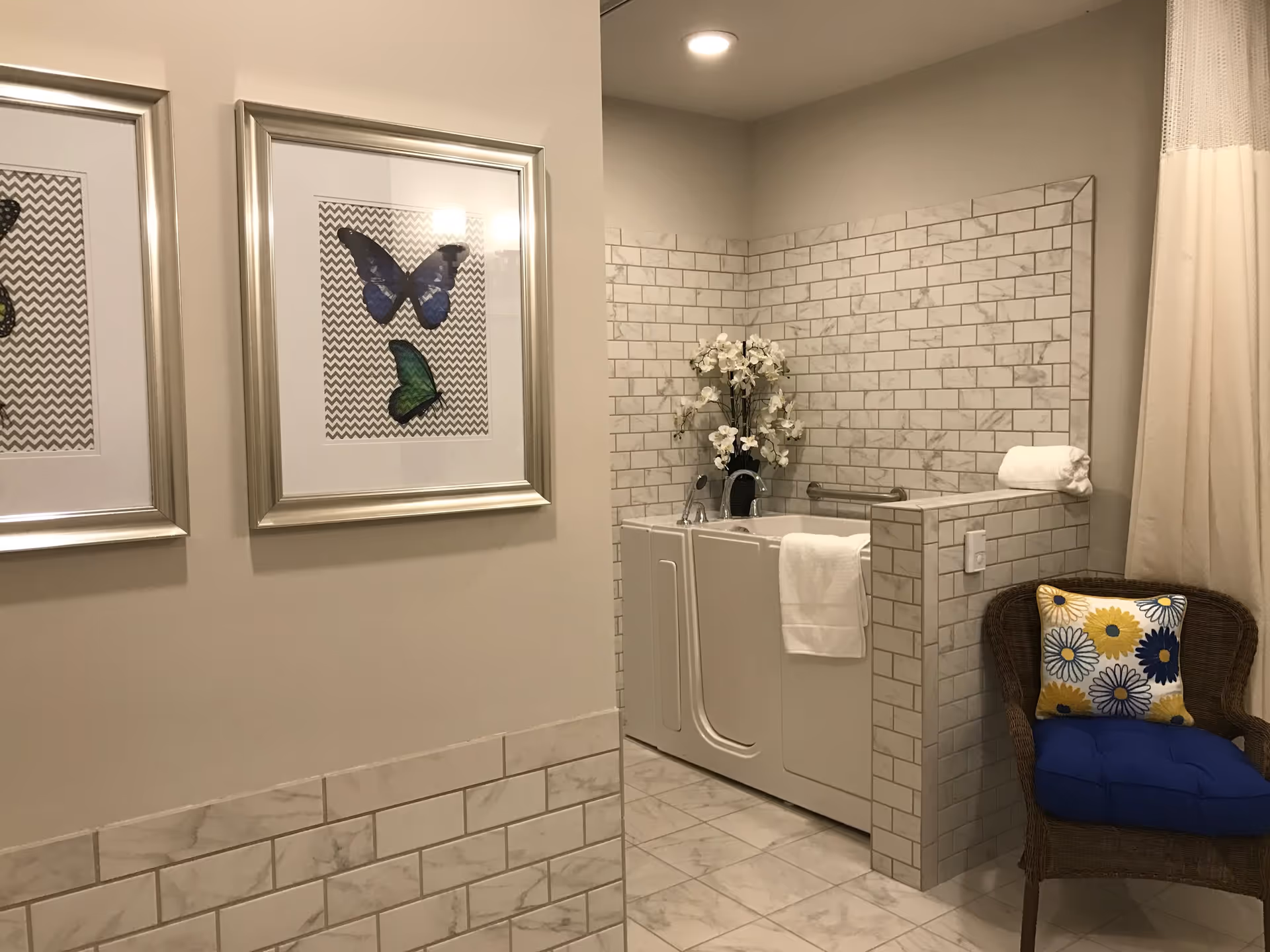 A bathroom featuring a walk-in bathtub with white tiled walls and floor. A white towel hangs on the tub, and a vase with white flowers is placed on the tub ledge. To the right, there is a wicker chair with a blue cushion and a decorative pillow with yellow and blue flowers. On the left wall, two framed butterfly artworks are displayed.