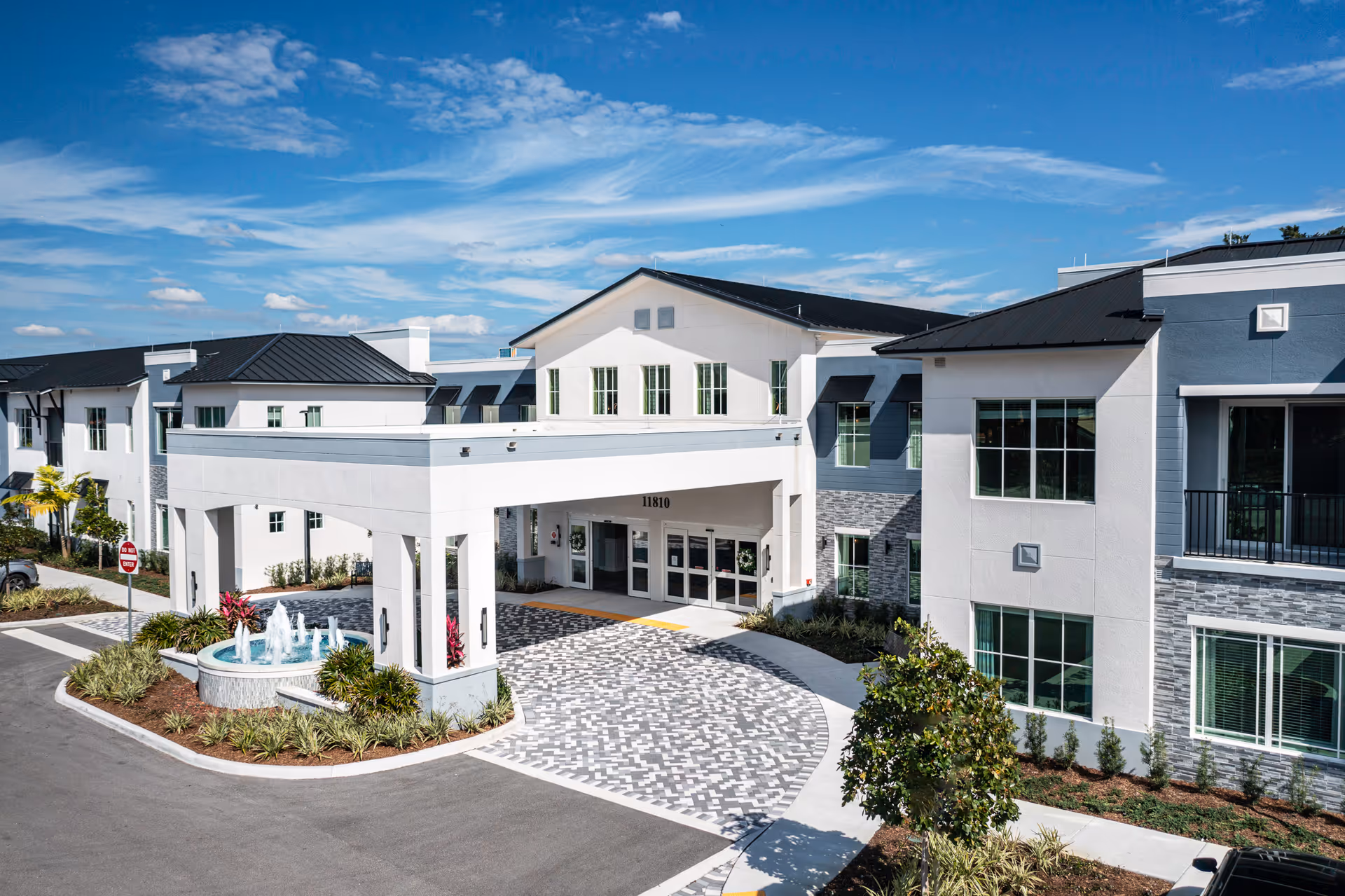 Front exterior view of HarborChase of Boynton Beach facility with a covered entrance, a circular driveway with patterned paving, a water fountain surrounded by landscaping, and a clear blue sky with scattered clouds.