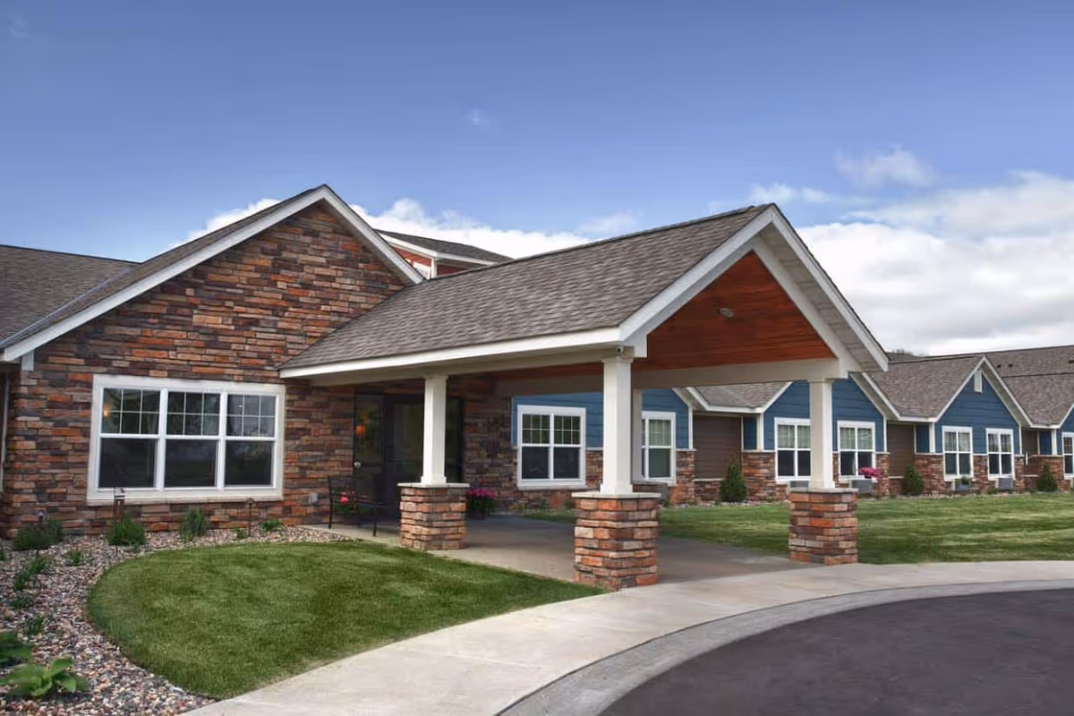 Exterior view of a single-story senior living facility building with a covered entrance supported by stone pillars. The building has a combination of brick and blue siding with multiple windows and a well-maintained lawn and landscaping under a partly cloudy sky.