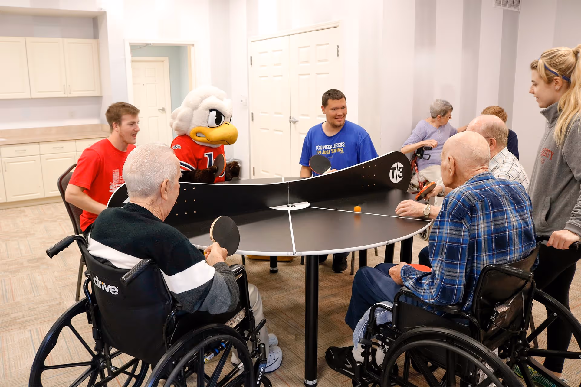 A group of elderly people and younger adults playing a game of table tennis on a round table with a net in the middle. Two elderly men in wheelchairs are holding paddles, while others, including a person in a bird mascot costume, watch and participate. The setting is a bright room with white walls and cabinets in the background.
