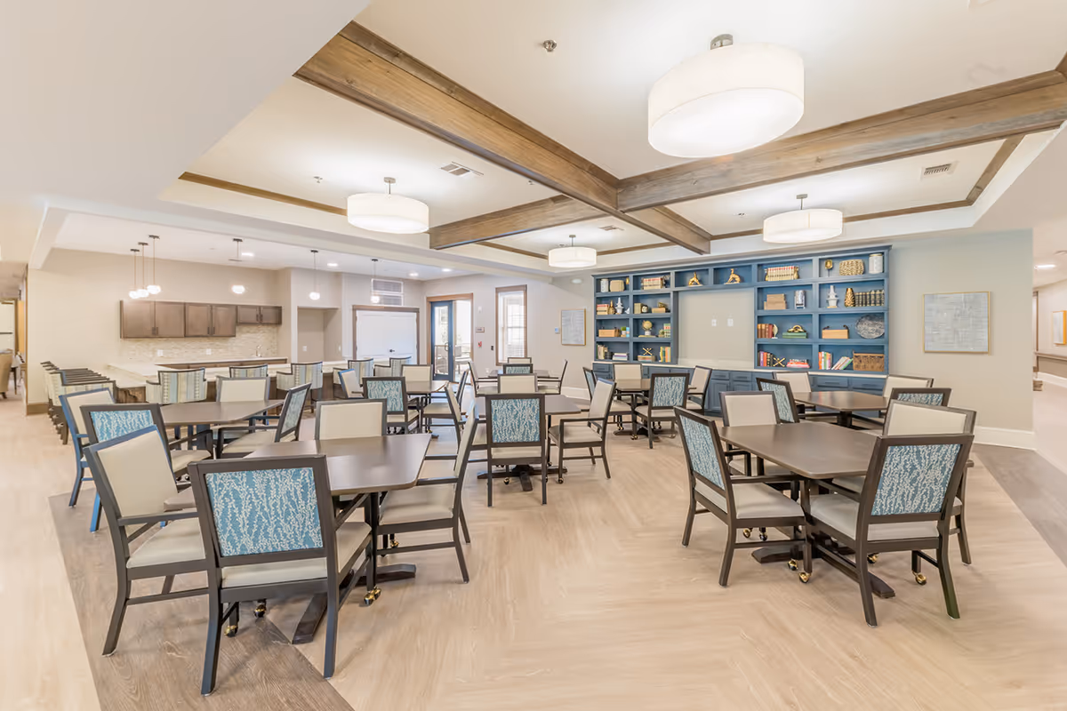 Spacious senior living dining room with multiple tables and chairs, pendant lights, and a blue built-in bookshelf.