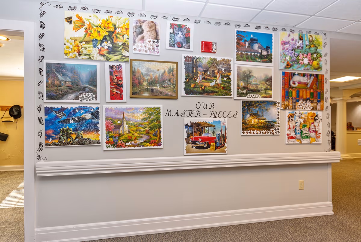 A wall display titled 'Our Master Pieces' featuring multiple framed jigsaw puzzles with various images including flowers, animals, landscapes, and vintage scenes, arranged neatly on a white wall in a well-lit hallway with carpeted floor.