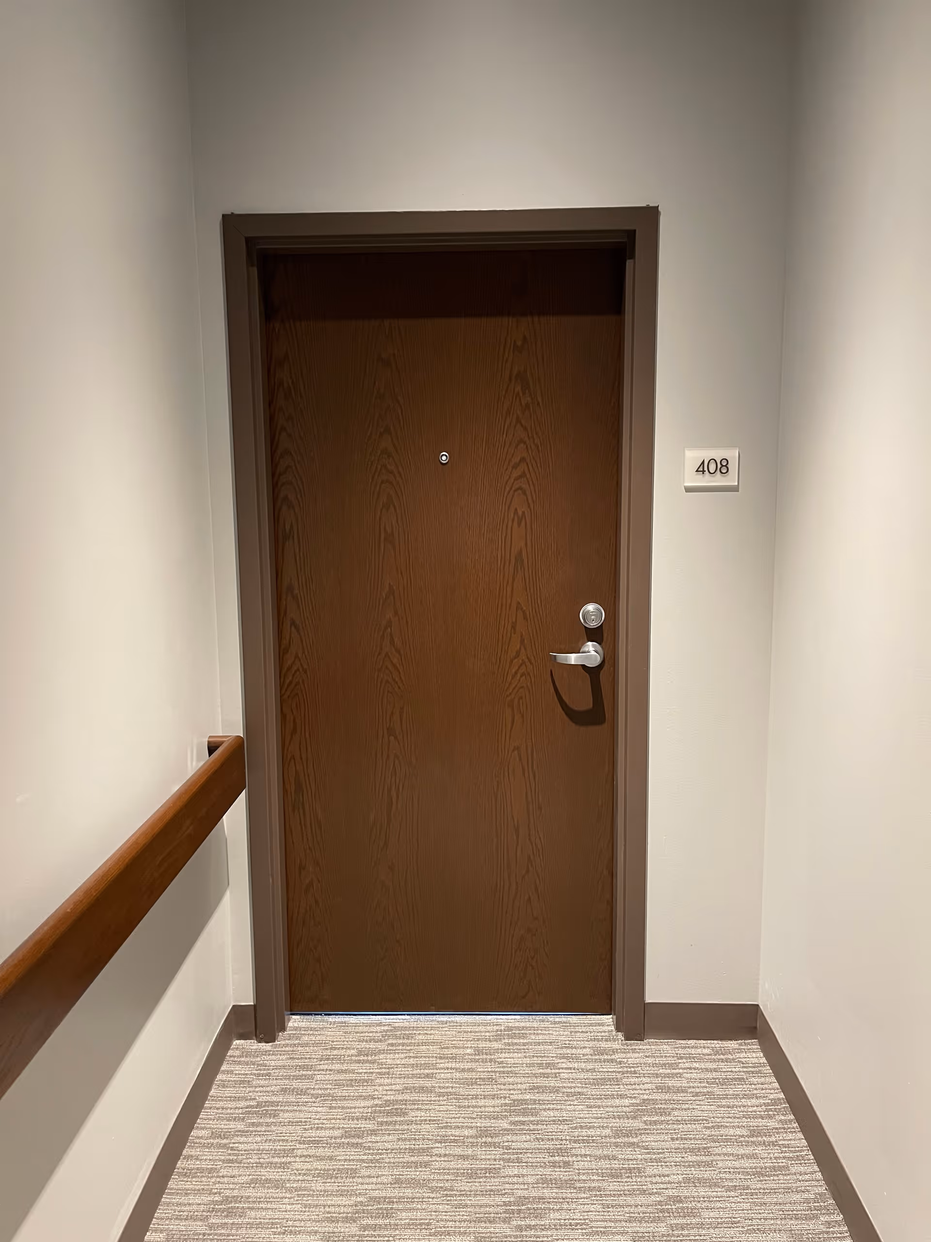 A closed wooden door with a silver handle and a peephole, set in a beige wall with a room number plaque labeled 408 to the right. There is a wooden handrail attached to the left wall and a carpeted floor in front of the door.