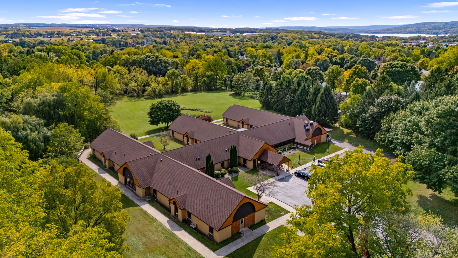 Aerial view of Clinton Crest Manor, a single-story building complex surrounded by lush green trees and open grassy areas under a clear blue sky.