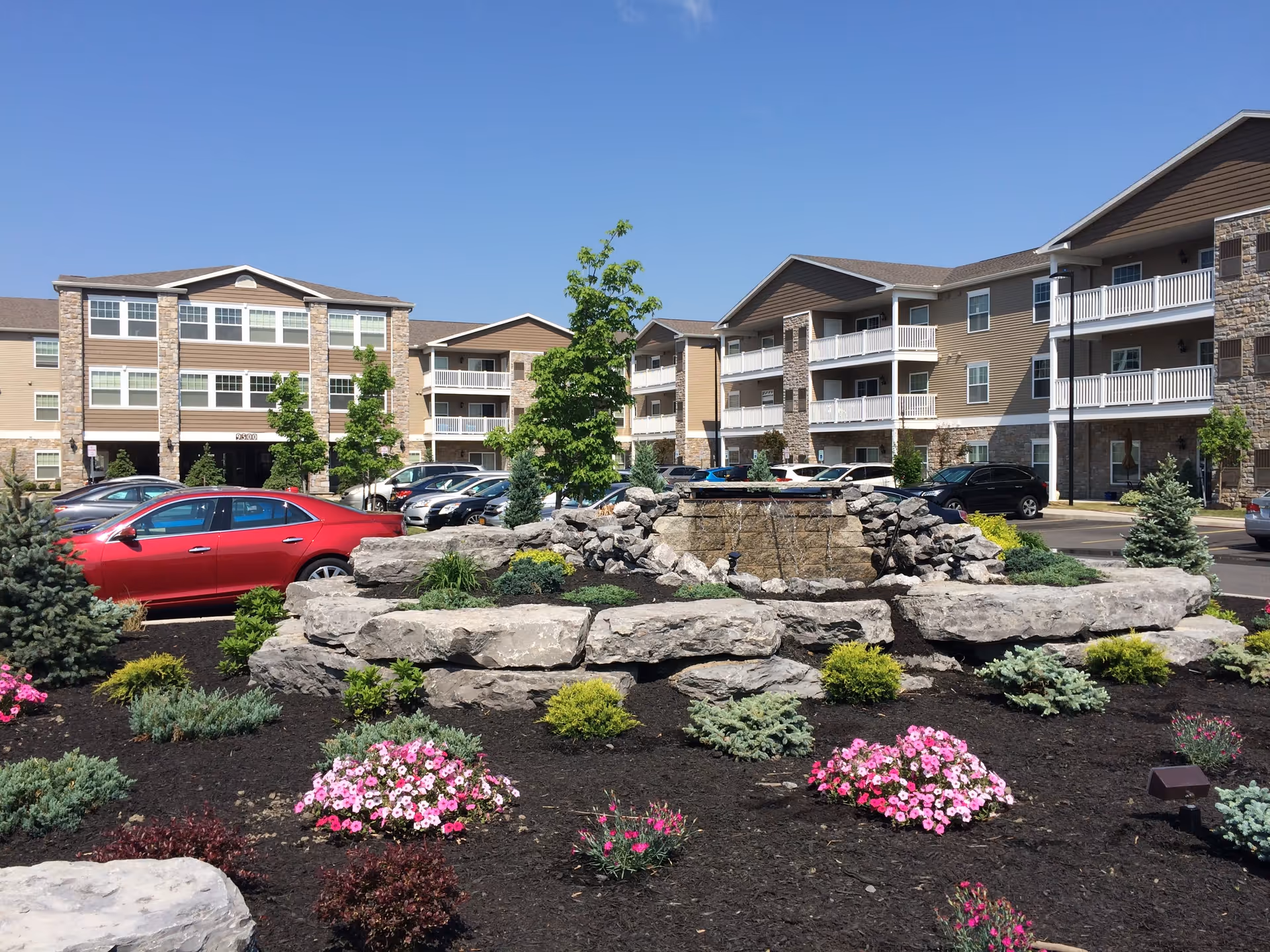 Outdoor view of Fox Creek Estates showing a landscaped garden with a rock water feature surrounded by various shrubs and pink flowers. Behind the garden, there is a parking lot with several cars and a three-story residential building with balconies and stone accents under a clear blue sky.