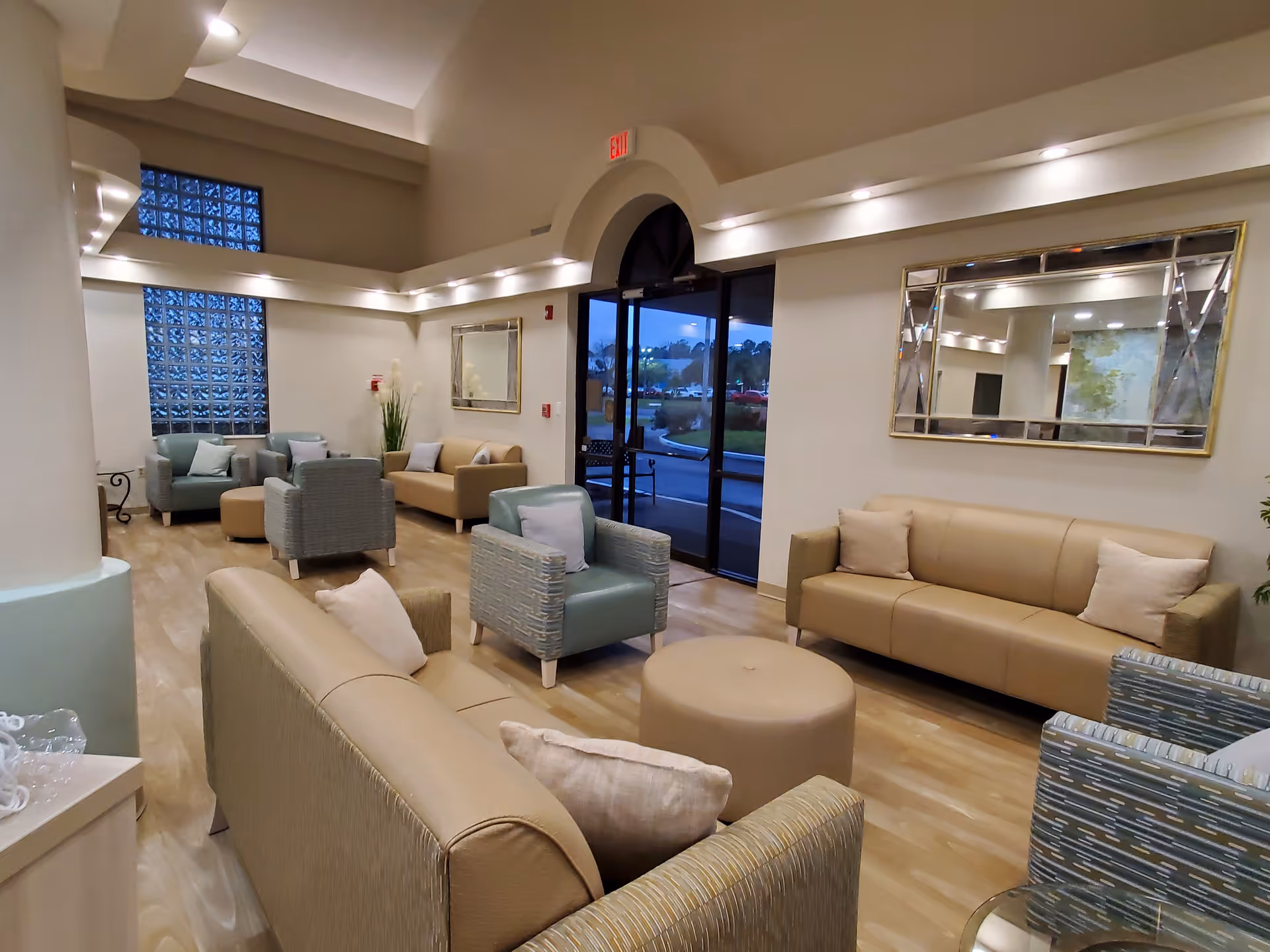 A spacious and well-lit assisted living facility common area with beige and light blue sofas and armchairs arranged around a round beige ottoman. The room features light wood flooring, a large decorative mirror on the wall, glass block windows, and a glass door leading outside. Soft recessed lighting highlights the high ceiling and neutral-colored walls.