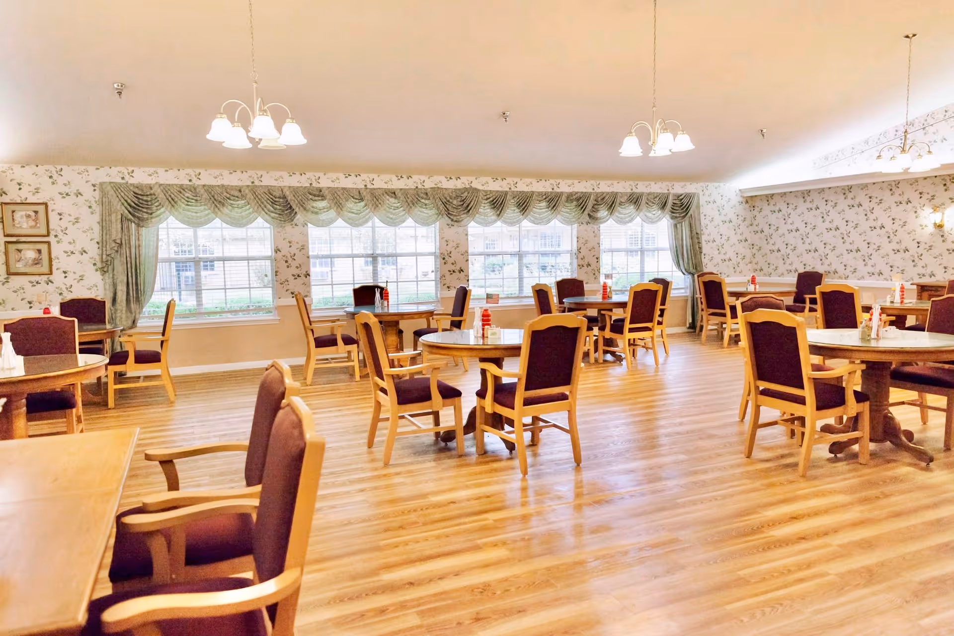 A spacious dining room with multiple round wooden tables and chairs with purple cushions. The room has large windows with green valance curtains, floral wallpaper, and wooden flooring. Several light fixtures hang from the ceiling, illuminating the room.