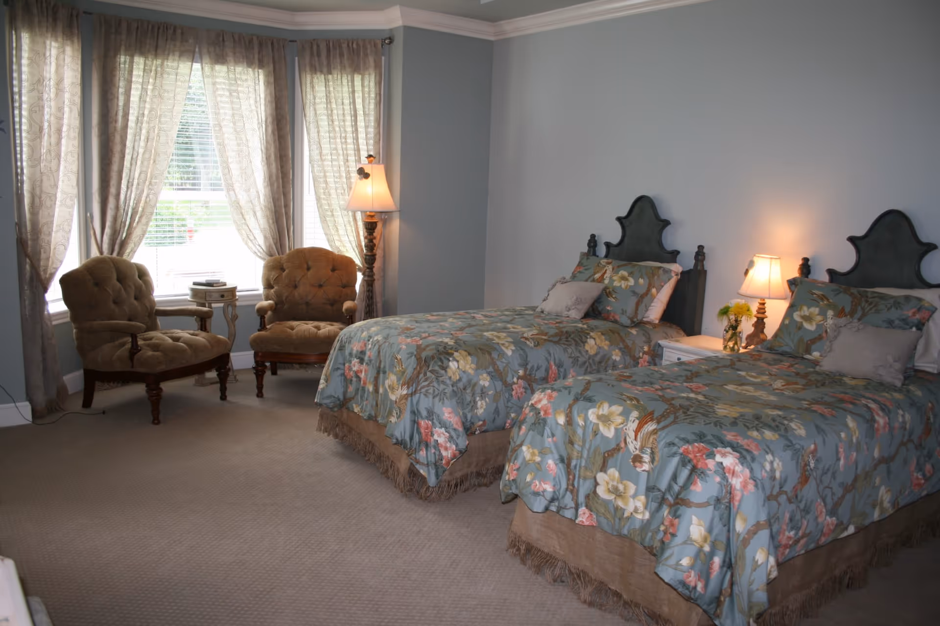 A bedroom with two twin beds featuring floral bedspreads, a nightstand with a lamp and flowers between the beds, two upholstered armchairs near a window with sheer curtains, and a floor lamp in the corner.