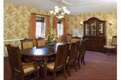 A traditional dining room with a long wooden dining table surrounded by eight cushioned chairs. The room features floral wallpaper, a chandelier hanging above the table, two windows with red curtains, a large wooden china cabinet, and a floral arrangement centerpiece on the table.