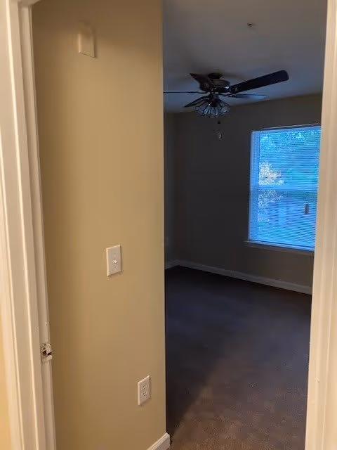 View through a doorway into a dimly lit bedroom with a ceiling fan and a window with blinds partially open showing some greenery outside. The walls are beige and the floor is carpeted.